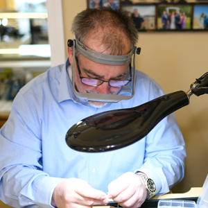 Leo Poisson closely examining a piece of jewelry with a jeweler's loupe on his head and a lamp beside him.