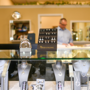 Earrings, necklaces, rings, and bracelets are delicately displayed on shelves with busts and cushions holding them, all in dark colors, with Leo Poisson in the background working on other shelves.