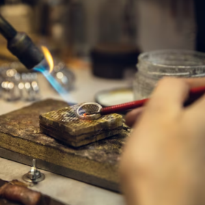 A ring placed on a wooden board on a workbench, being resized with a jeweler's torch that melts the metal with fire.