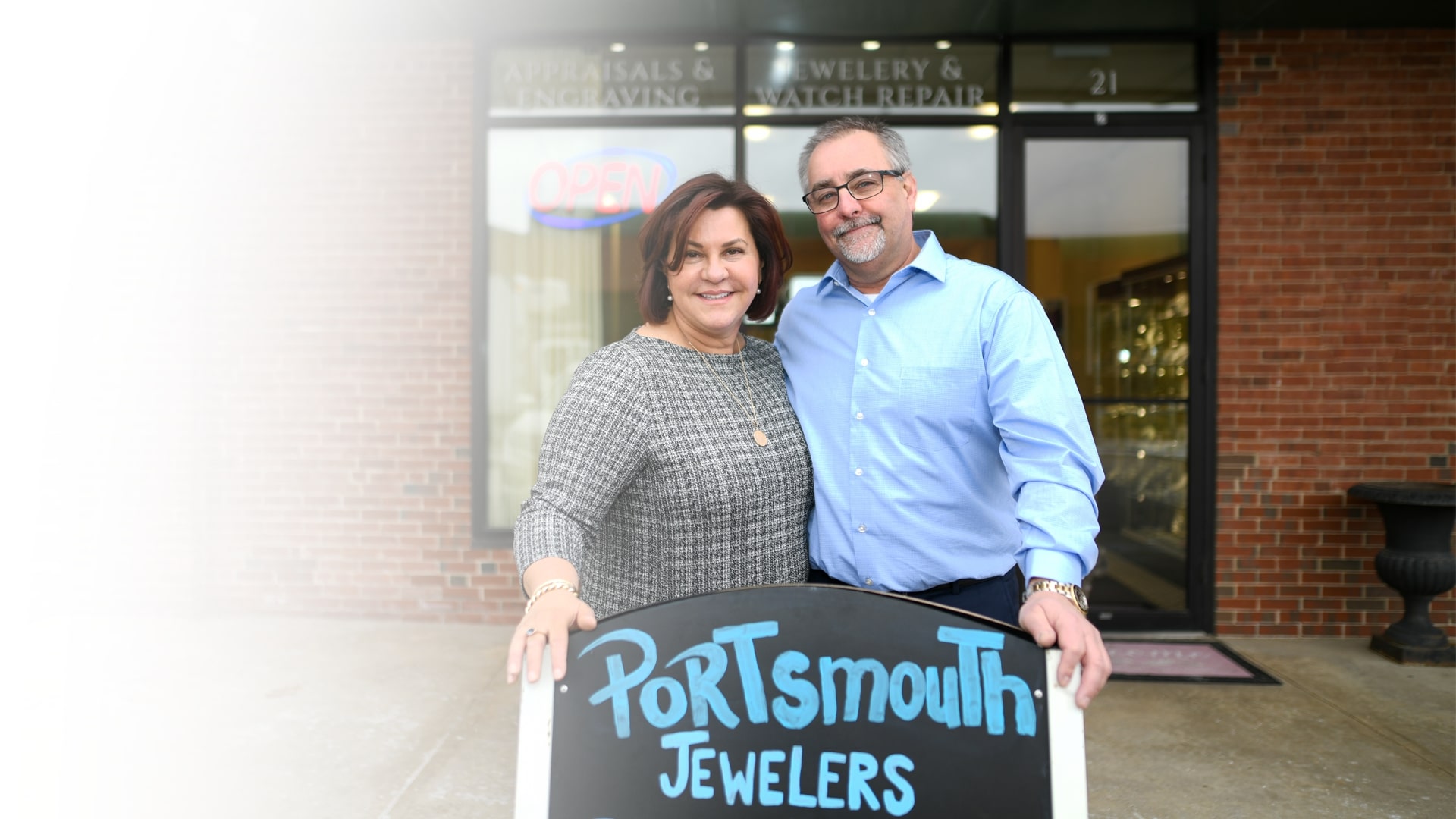 From left to right, Lisa Poisson, wearing a tweed suit and jewelry, and Leo Poisson, wearing a light navy blue shirt, dress pants, and a watch, smiling as they pose in front of the entrance to Portsmouth Jewelers.
